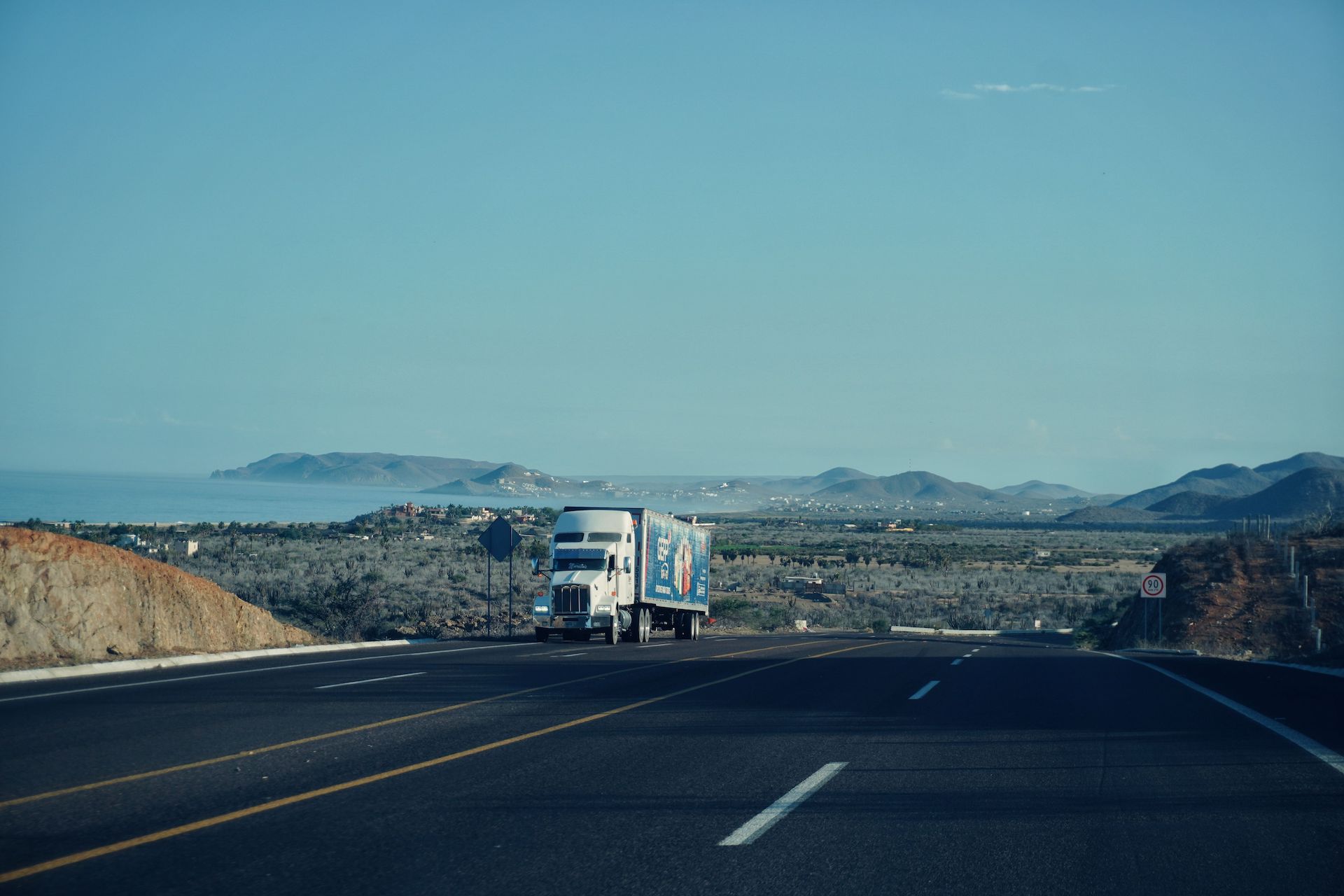 Trailer Transit, Inc. | A white semi-truck drives on a highway with mountains and a coastal landscape in the background under a clear blue sky.