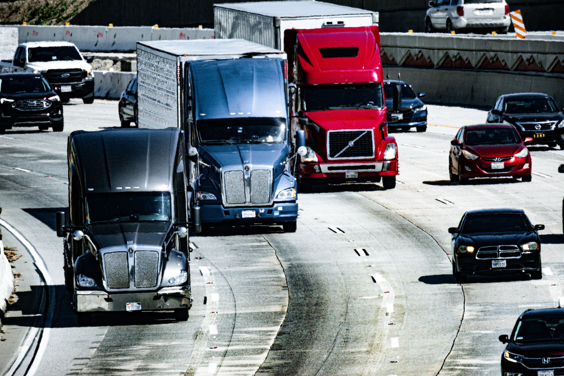 Trailer Transit, Inc. | Several large semi trucks and cars drive on a multi-lane highway bordered by concrete barriers on a clear day.