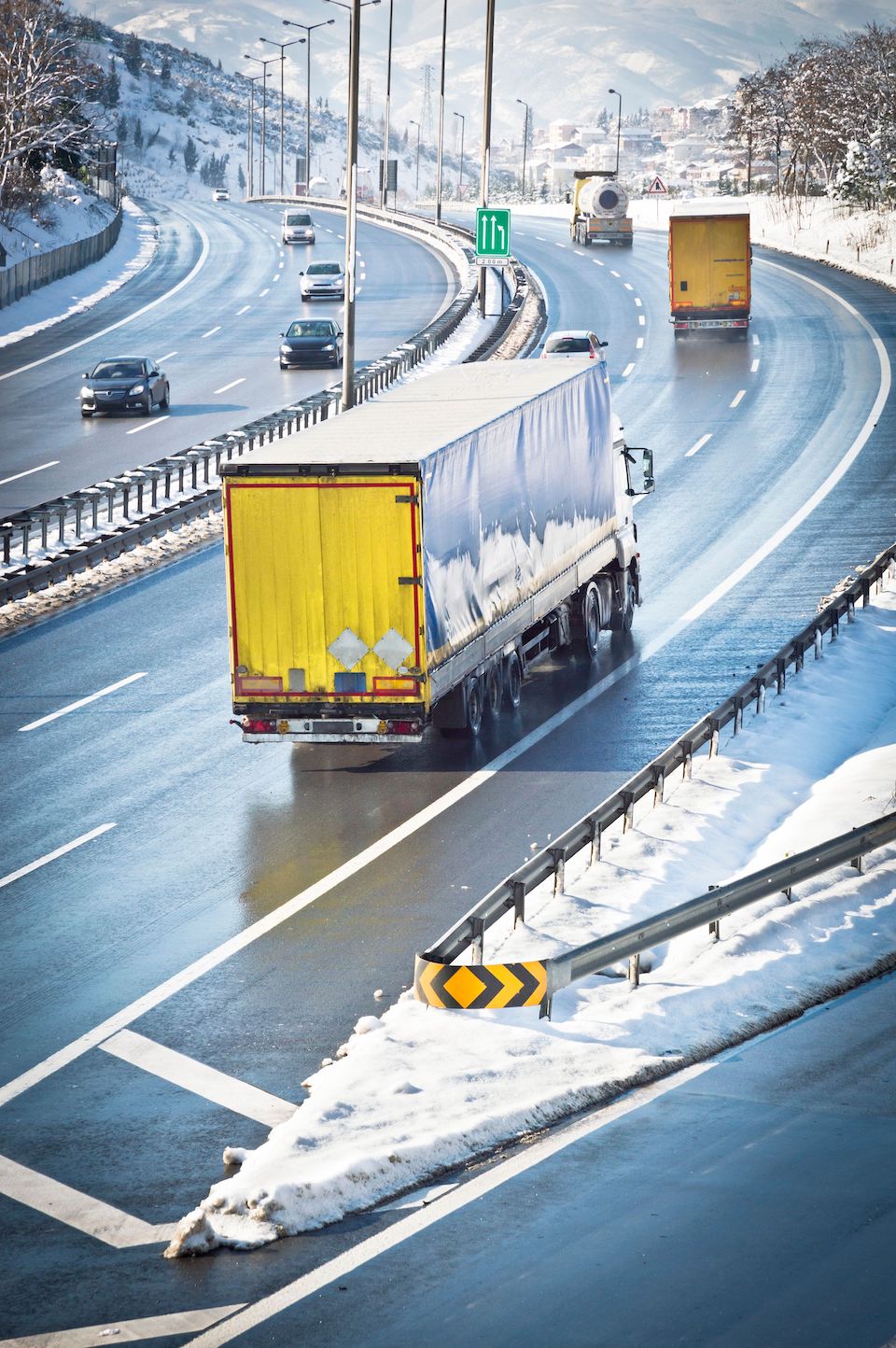 Trailer Transit, Inc. | A yellow truck drives on a wet, snow-lined highway with several other vehicles. Snow covers the road’s edges and the truck’s roof. Road signs and mountains are visible in the background.