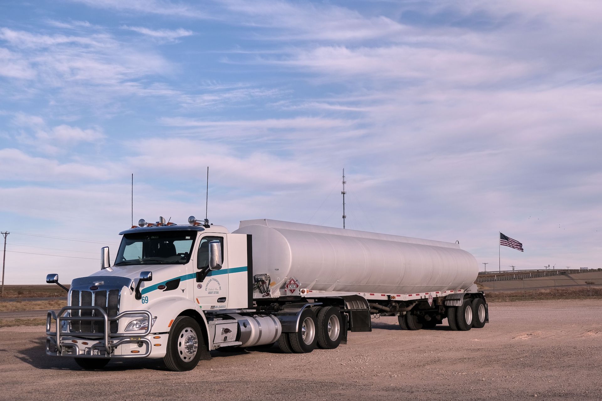 Trailer Transit, Inc. | White tanker truck parked on a gravel lot with a US flag and radio tower in the background under a partly cloudy sky.