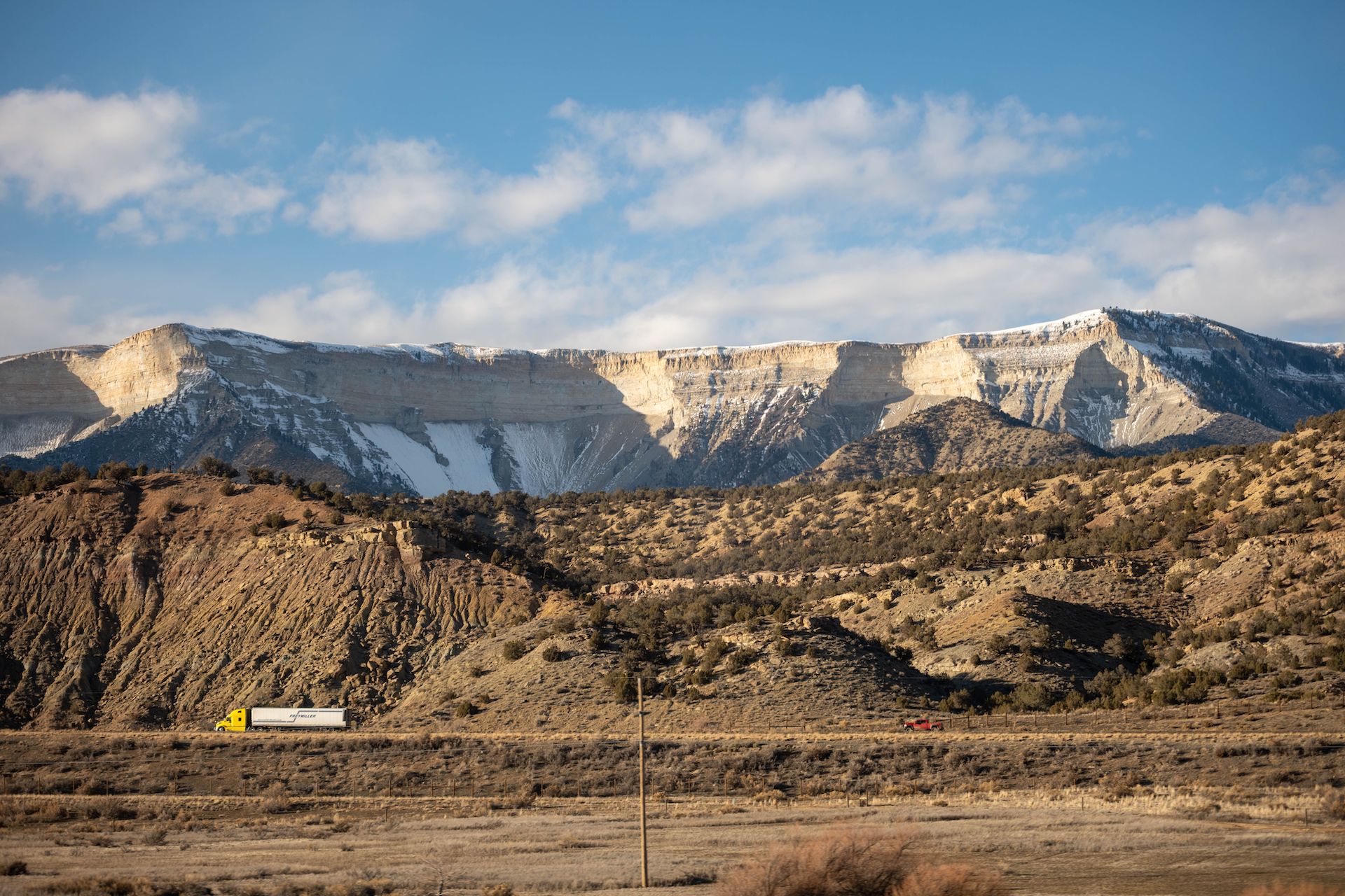 Trailer Transit, Inc. | A yellow semi-truck drives along a highway through a dry, hilly landscape with snow-capped mesas in the background under a partly cloudy sky.