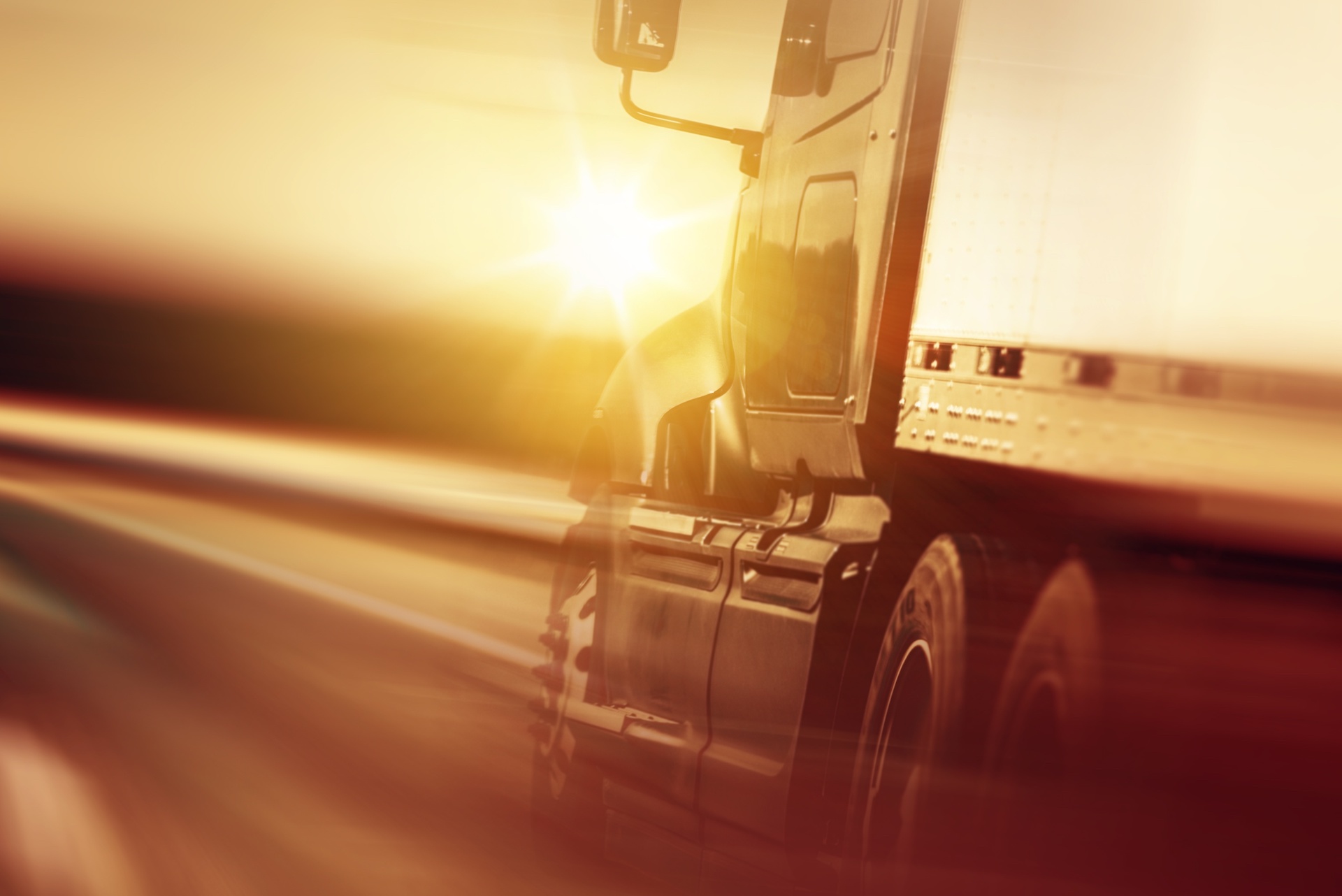 Trailer Transit, Inc. | Close-up of a semi-truck driving on a highway at sunset, with motion blur emphasizing speed and sunlight reflecting off the vehicle.