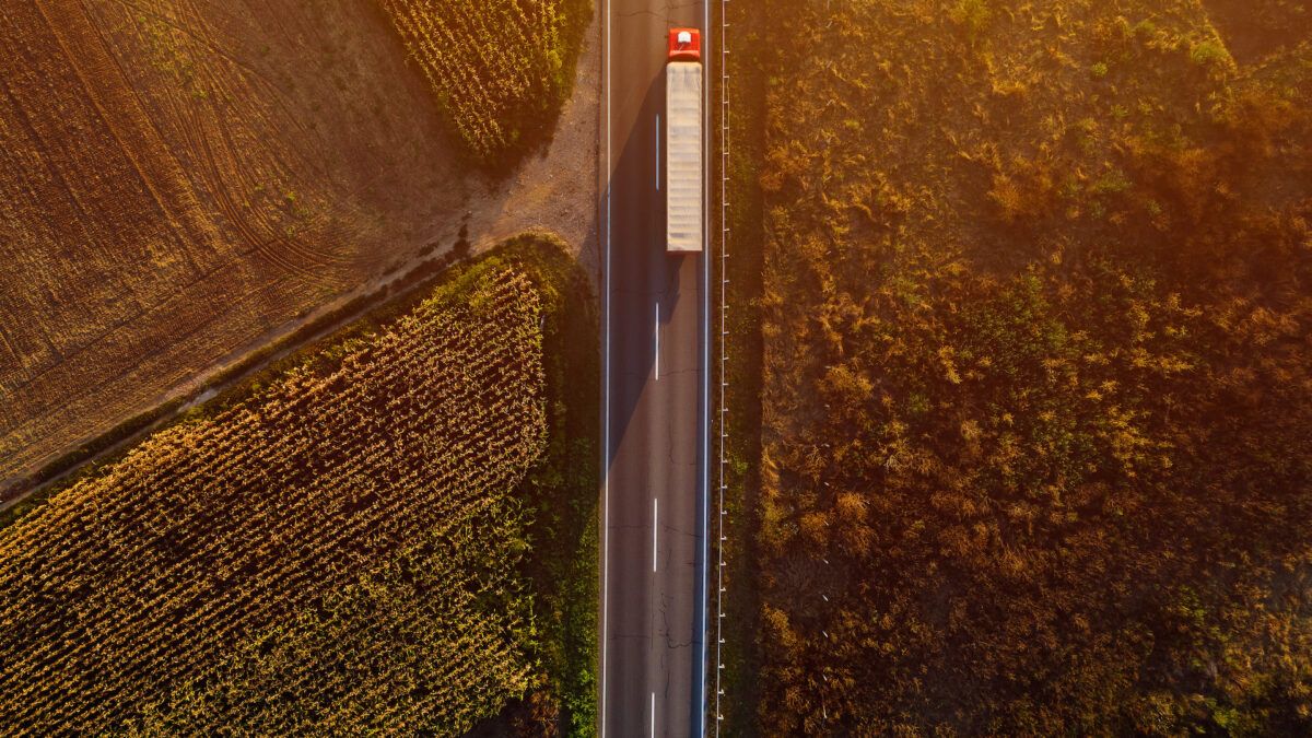 Trailer Transit, Inc. | An aerial view of a long-haul truck driving on a straight rural road, flanked by agricultural fields during sunset.
