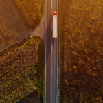 Trailer Transit, Inc. | An aerial view of a long-haul truck driving on a straight rural road, flanked by agricultural fields during sunset.