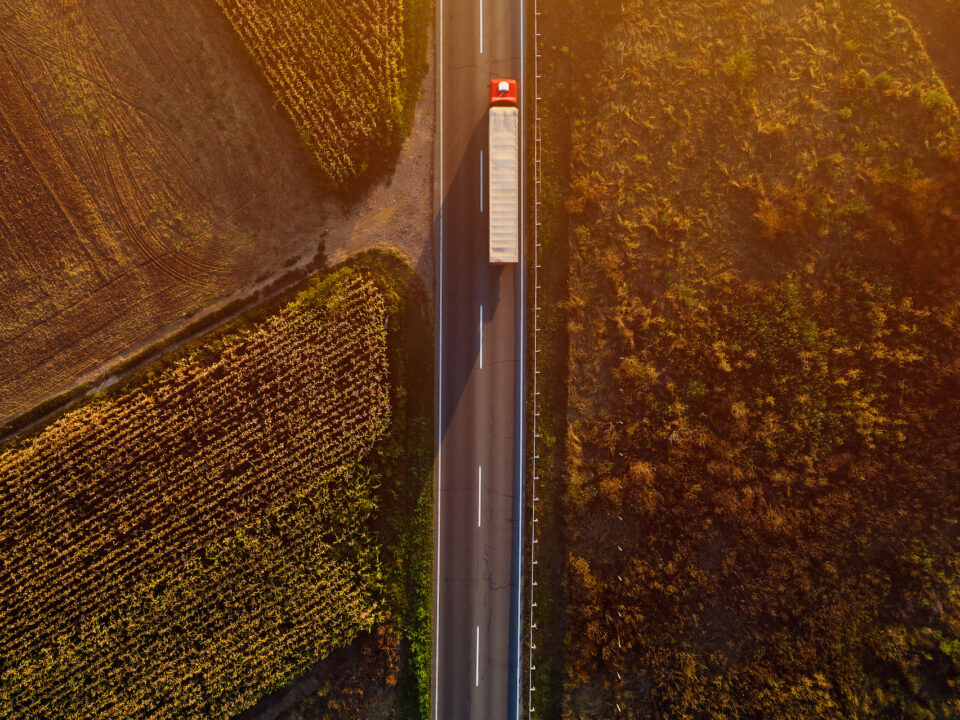 Trailer Transit, Inc. | An aerial view of a long-haul truck driving on a straight rural road, flanked by agricultural fields during sunset.