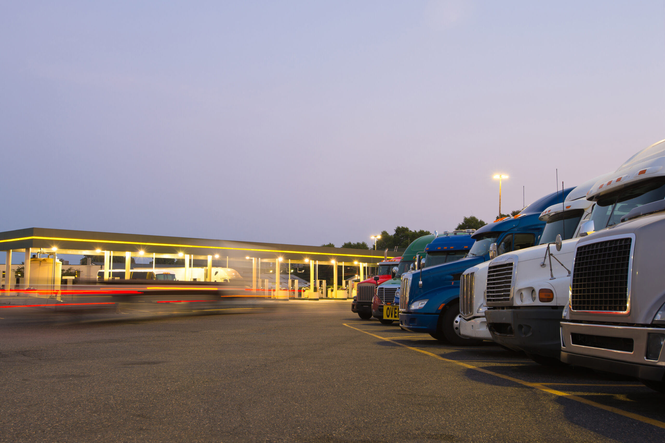 Trailer Transit, Inc. | Several semi-trucks parked in a row at a truck stop during dusk, with blurred vehicle lights passing by in the background.
