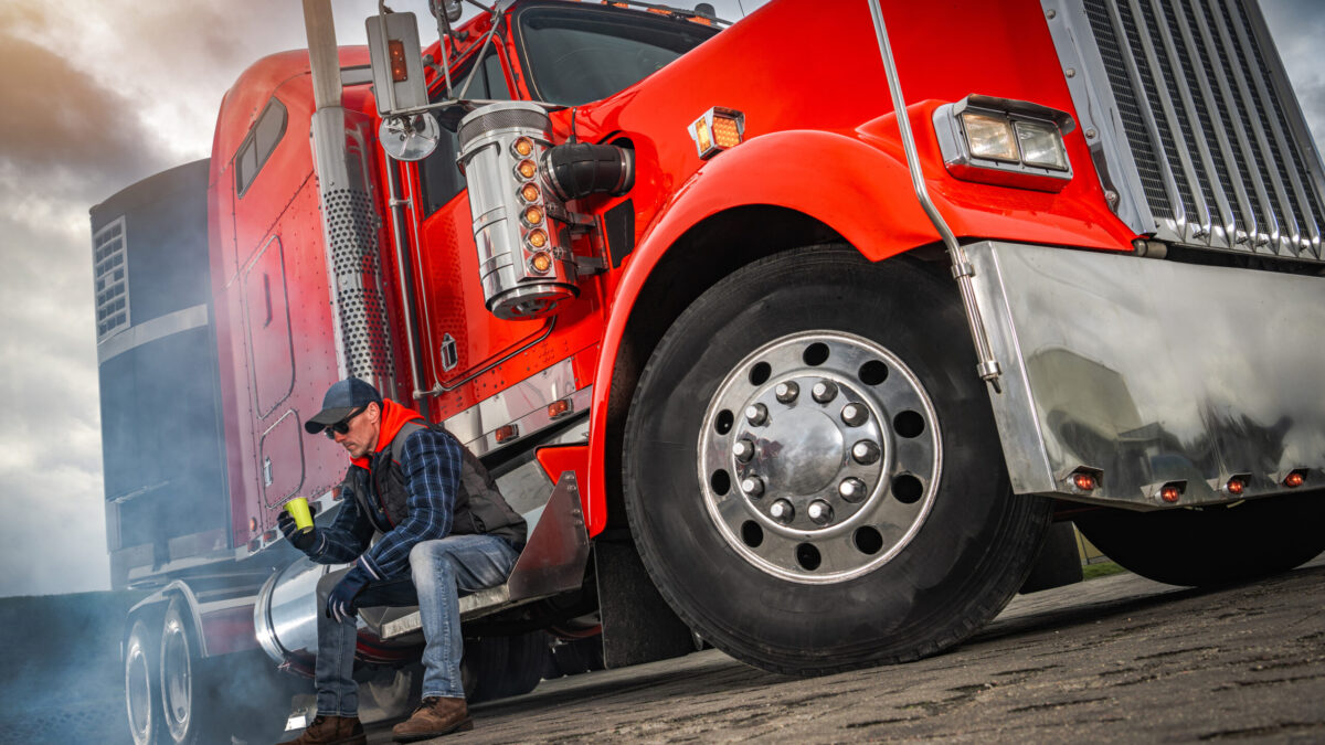 Trailer Transit, Inc. | A truck driver wearing a hat and sunglasses sits on the step of a red semi-truck, having a coffee break.