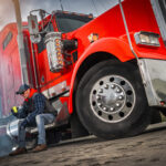 Trailer Transit, Inc. | A truck driver wearing a hat and sunglasses sits on the step of a red semi-truck, having a coffee break.