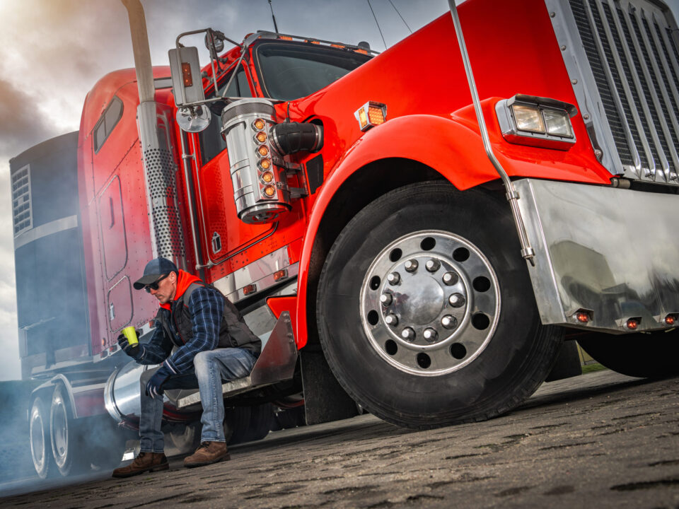 Trailer Transit, Inc. | A truck driver wearing a hat and sunglasses sits on the step of a red semi-truck, having a coffee break.