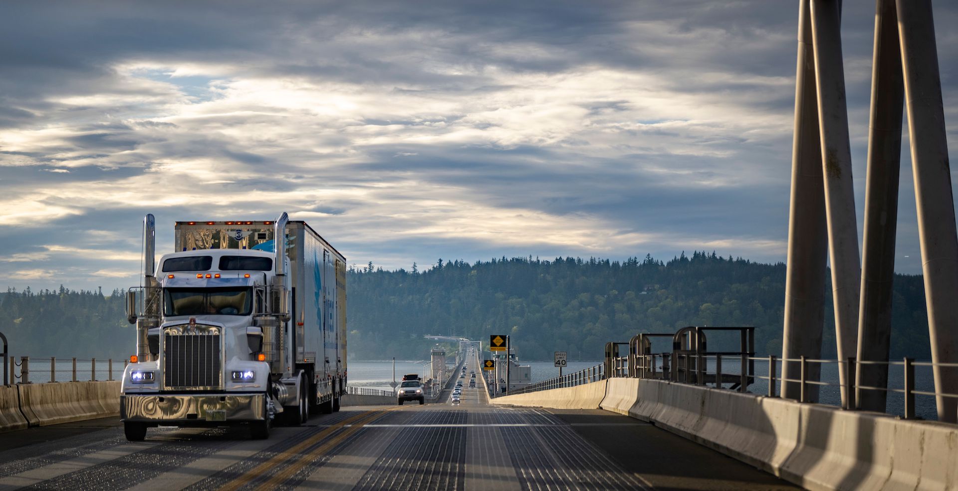 Trailer Transit, Inc. | A semi-truck drives on a long bridge over water, with a line of cars following in the distance.