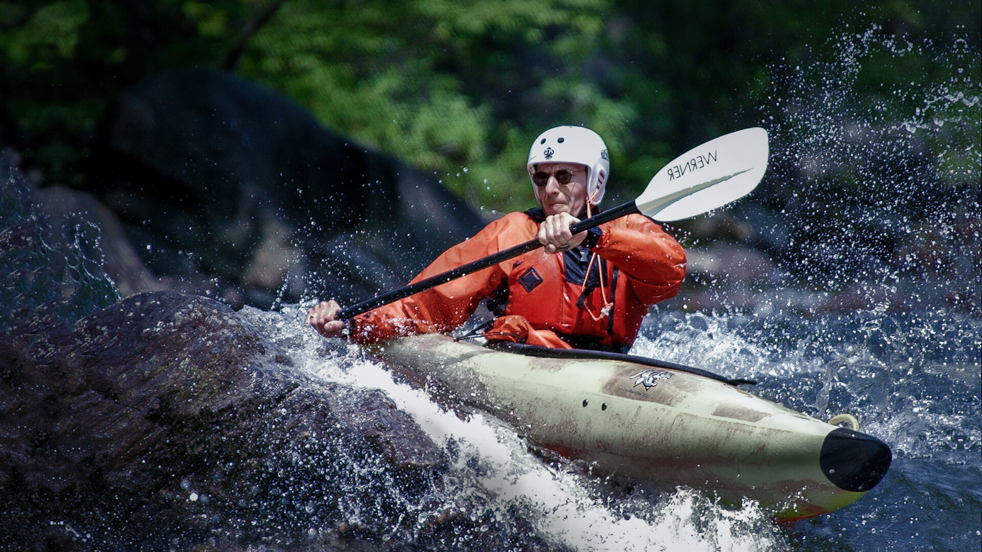 Trailer Transit, Inc. | Person in an orange jacket and white helmet kayaking through rough, rocky river rapids, water splashing around.